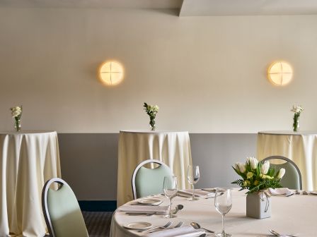 Banquet setup with white tablecloths, glassware, floral centerpieces, and two warm wall lights in a bright event room.