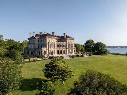 A grand white mansion with red roof stands on a manicured lawn by the water under a clear blue sky.