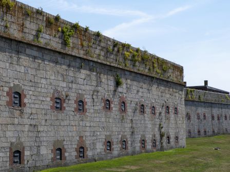A long gray stone fort wall with red-brick gun embrasures, overgrown with ivy, under a blue sky.
