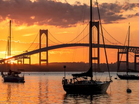 A suspension bridge towers at sunset over calm water, with sailboats anchored in the foreground and golden clouds above.