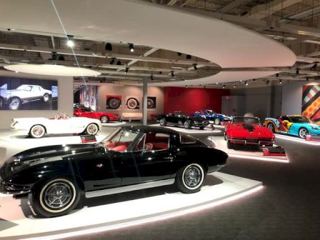 A car museum showroom filled with classic sports cars, including a black convertible and a red Corvette under bright lights.