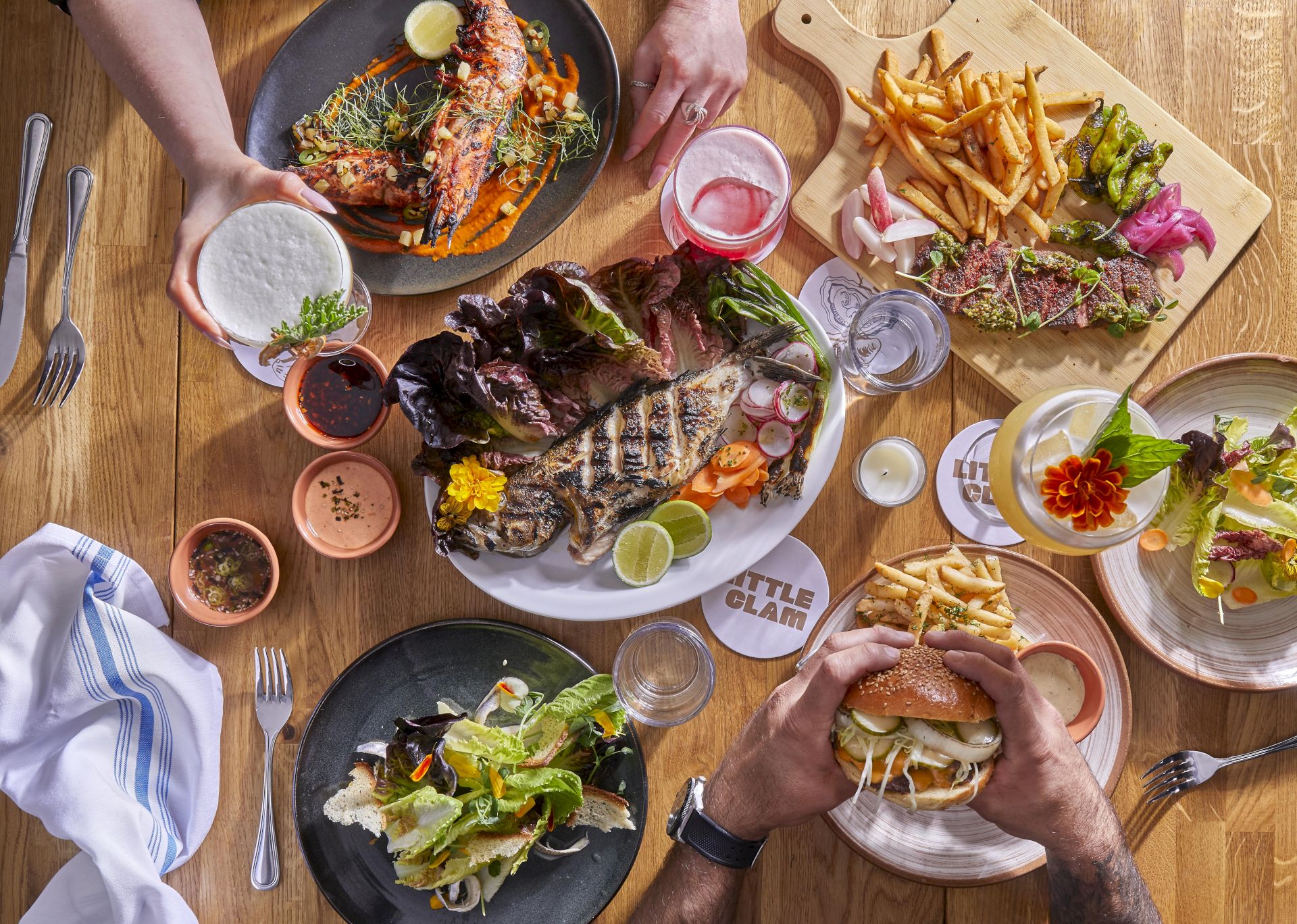 A table full of varied dishes: burgers, fries, salads, sauces, and drinks shared among several people at a wooden table.