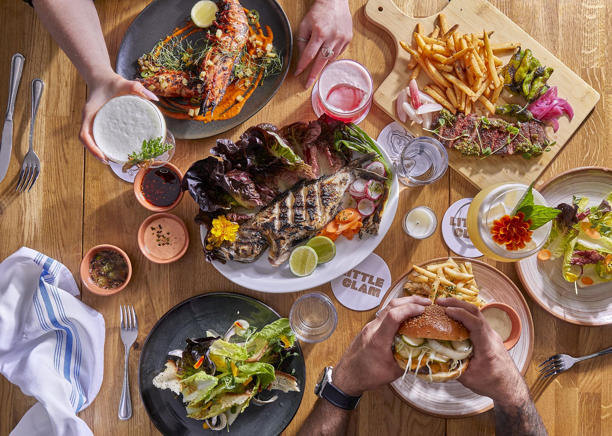 A table full of varied dishes: burgers, fries, salads, sauces, and drinks shared among several people at a wooden table.