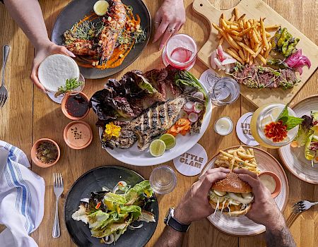 A top-down spread of a multi-dish meal: grilled meats, fries, salads, seafood, and drinks on wooden tables.