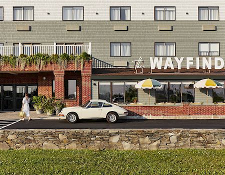A hotel exterior with the “WAYFINDER” sign, a parked white classic car, patio umbrellas, and a woman walking by.