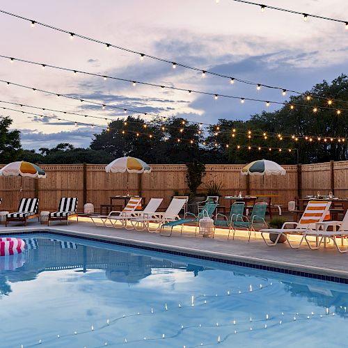 A swimming pool is surrounded by lounge chairs, umbrellas, and string lights in a cozy outdoor setting. The sky is partially cloudy at dusk.