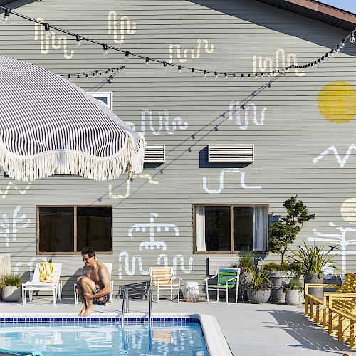 A pool area with lounge chairs, a person sitting by the pool, and a building with colorful wall art and string lights in the background.