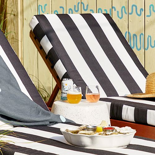 Two lounge chairs with black and white stripes, drinks, and a straw hat on a table next to a bowl of oysters.