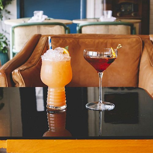 Two cocktails on a black table: one in a short glass filled with ice, the other in a tall martini glass. Leather chairs in the background.