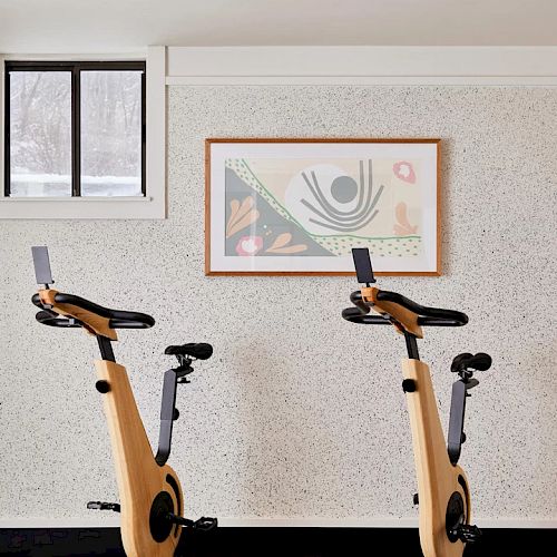 The image shows two stationary bikes in a room with a textured wall, a framed abstract artwork, and a small window.