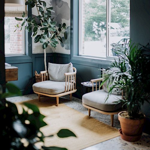 A cozy corner with two wooden chairs, cushions, a side table, large plants, and big windows letting in natural light, promoting a serene atmosphere.