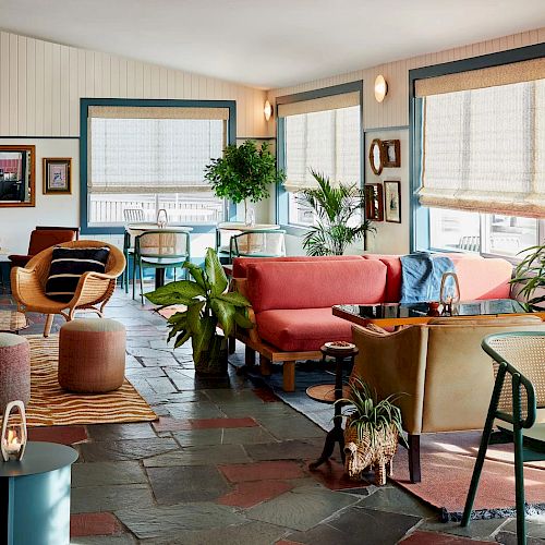 A cozy, eclectic living space with colorful furniture, plants, art on the walls, and a mix of wood and stone elements under natural light.