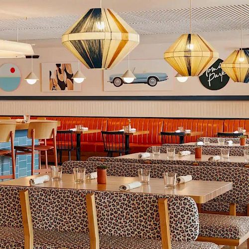 A modern restaurant with patterned seating, wooden tables, artistic decor, and stylish hanging lights. The walls feature various artistic pictures.