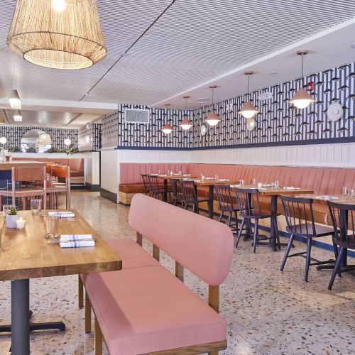 A stylish, airy restaurant with pink upholstered benches, wooden tables, modern pendant lights, and geometric blue-and-white wall accents, ready for dining.