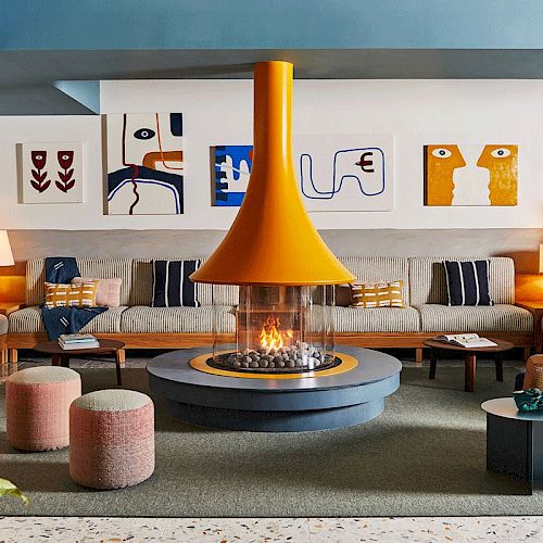 A modern living room features a central fireplace with a stylish orange hood, surrounded by sectional seating, abstract art, and cozy cushions.