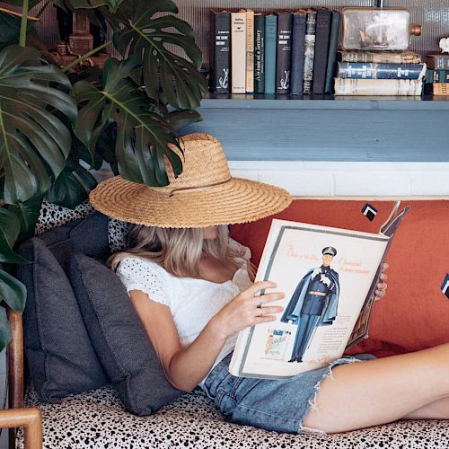 A person with a straw hat reads a book on a cozy couch surrounded by cushions, potted plants, and a bookshelf filled with books, creating a relaxed vibe.