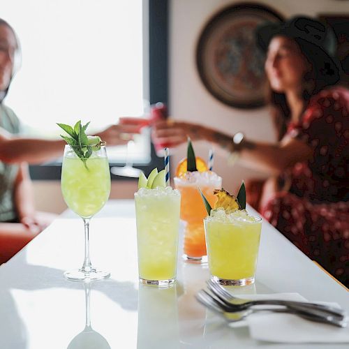 Two people are sitting at a table with colorful drinks in focus, with some silverware and napkins in the foreground.