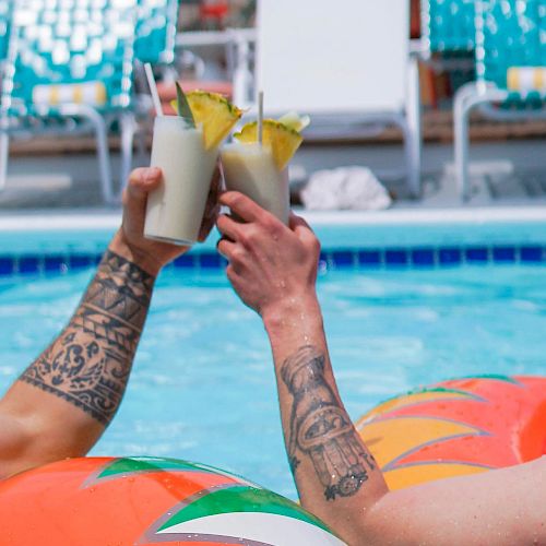 Two people clink drinks garnished with pineapple while floating in a pool on inflatable tubes.