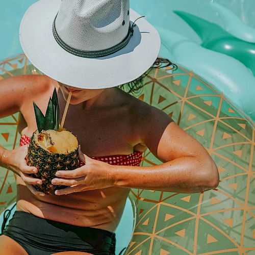 A person wearing a hat and swimsuit is relaxing on a pool float with a drink in a hollowed-out pineapple in a pool.