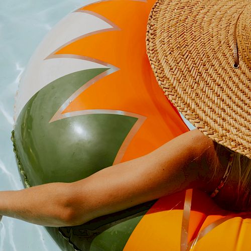 A person floats in a pool on a colorful inflatable while wearing a large straw hat, with one arm resting on the edge of the float.