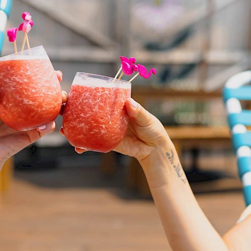 Two people are holding up glasses of a red slushy drink garnished with small pink flowers, sitting on blue and white chairs.