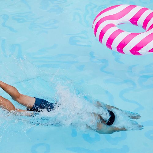 A person is diving into a swimming pool while a pink and white striped inflatable pool ring floats nearby in the water.