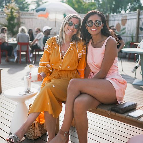 Two women sit close together outdoors, smiling at the camera, both wearing sunglasses, with other people and greenery in the background.