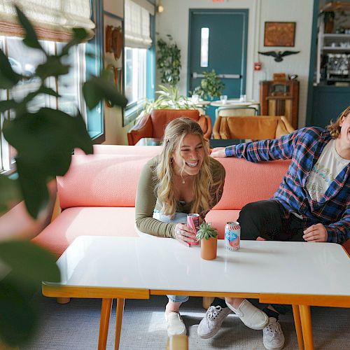 Two people are sitting on a pink couch, laughing, and holding drinks at a white table in a cozy, well-lit room with plants and a blue door.