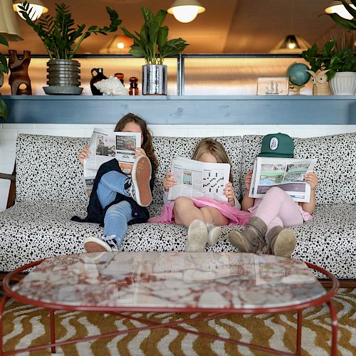 Three children sit on a couch, each holding and reading a newspaper, surrounded by plants and decor in a cozy living room setting.