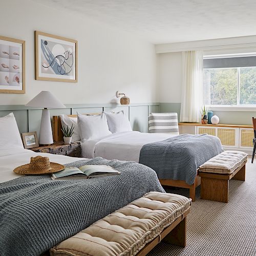 A bright, cozy room with two beds, framed art, a window, a small round table with a chair, and a straw hat on one of the beds ending the sentence.