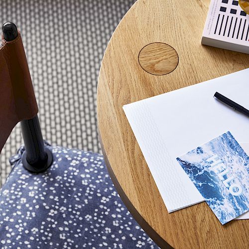 A small wooden table with a notebook, pen, postcard, and sunglasses sits next to a chair with a patterned cushion and tiled floor.