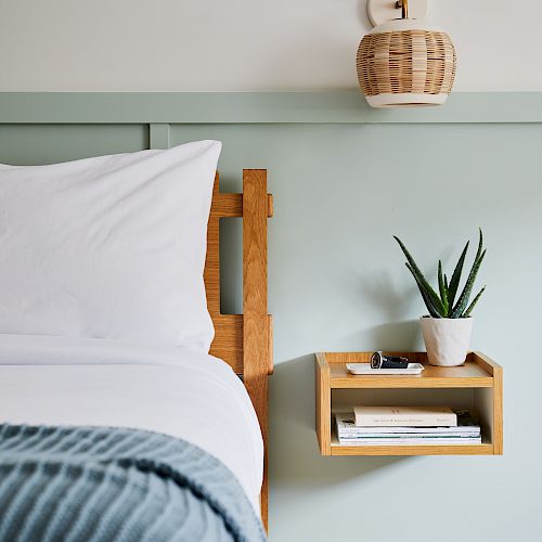 A neatly arranged bedroom corner with a bed, lamp, floating shelf holding a plant, glasses, two books, and a blue blanket.