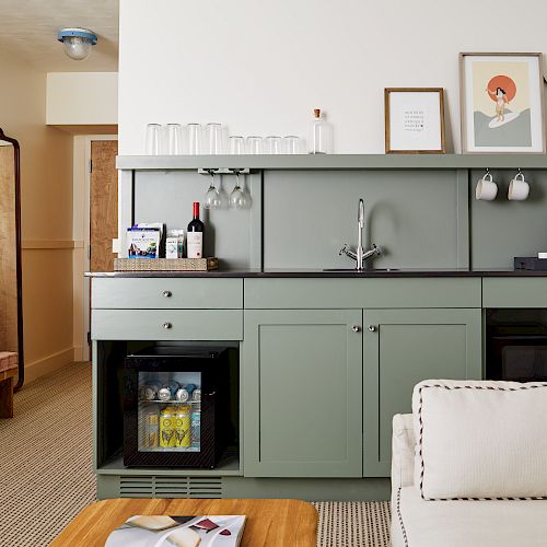 A cozy kitchenette with a mini-fridge, sink, coffee maker, and storage. Decor includes framed artwork, glasses, and potted plants.