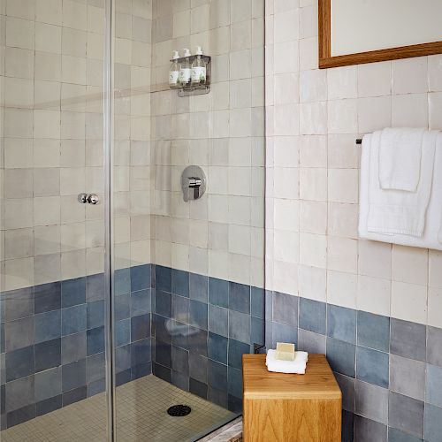 A modern bathroom with a glass-enclosed shower, wooden stool with soap and towel, and white towel on a wall rack.