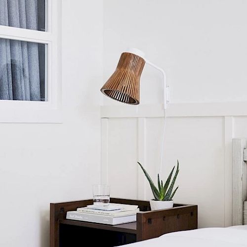 A minimalist bedroom features a bedside table with a plant, a glass of water, books, and a wooden lampshade against a white wall and window.