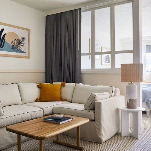 The image shows a modern living space with a beige sectional sofa, a wooden coffee table, a lamp, and decorative items with a partition to a bedroom area.