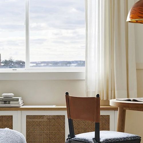 A cozy, well-lit room with a window view, a table, chair, books, a drink, and an orange pendant lamp.