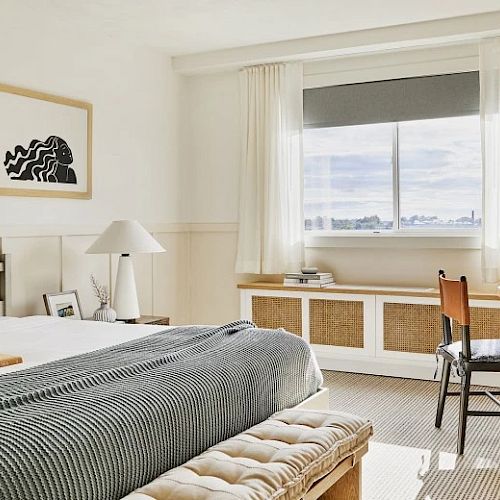 A cozy, modern bedroom featuring a bed with gray bedding, a wooden table and chair, wall art, and a large window with a scenic view, is well-lit.