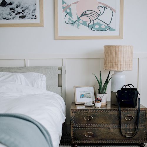 A bedroom with a bed, two framed artworks, a nightstand holding a lamp, plant, picture frame, and bag, and a pair of black socks on the floor.