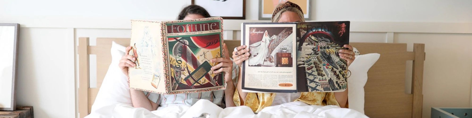 Two people are sitting up in bed reading magazines, with framed art on the wall behind them and a breakfast tray with a coffee pot and cup in front.