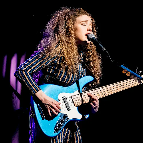 A curly-haired musician sings into a microphone while playing a bright blue electric guitar on stage.
