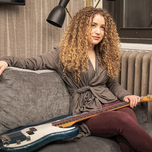 A curly-haired woman lounges on a couch holding an electric bass in a softly lit studio.