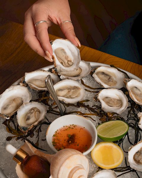 A person shucks fresh oysters over ice, with lemon wedges and a dipping sauce bowl on a platter.