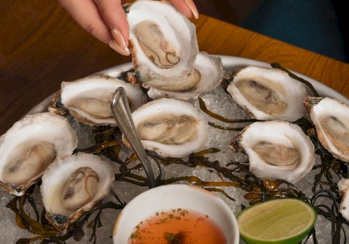 A person shucks fresh oysters over ice, with lemon wedges and a dipping sauce bowl on a platter.