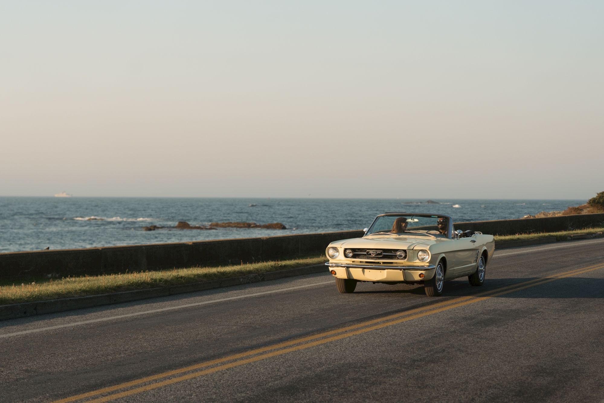 A white vintage convertible with two people drives along a coastal road beside the ocean at sunset.