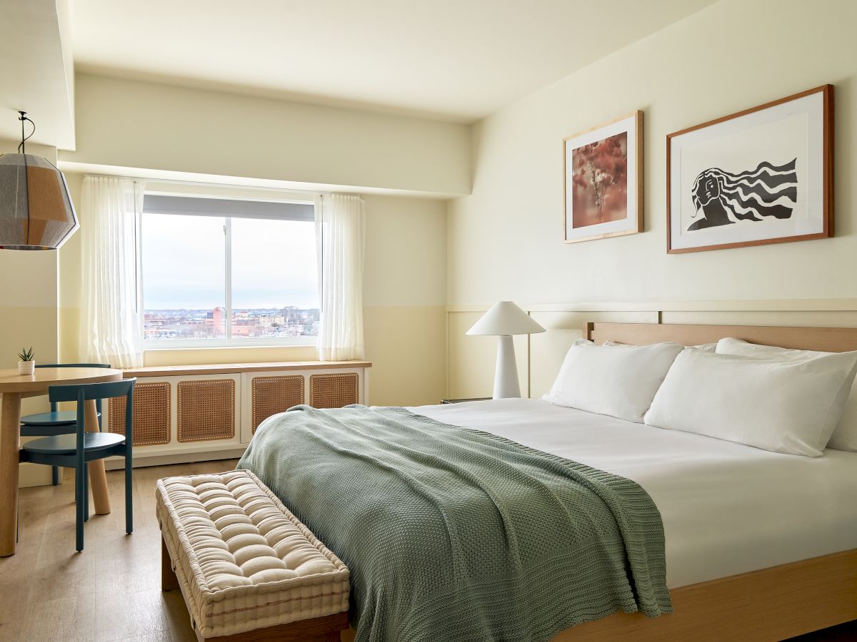 A bright modern bedroom with a large bed, bedside lamps, framed wall art, and a window with white curtains and city views.