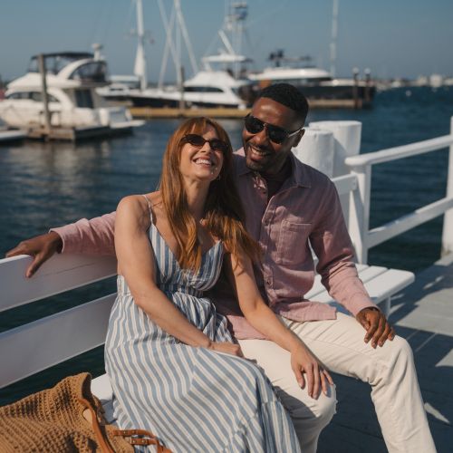 A smiling couple lounges on a yacht deck in sunny harbor, sunglasses on, with boats and docked ships in the background.