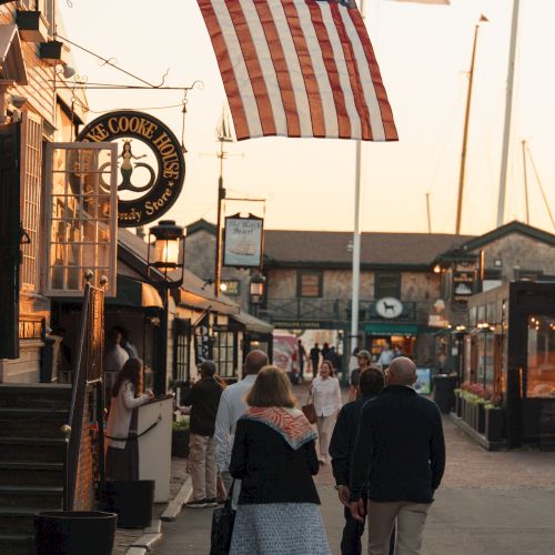 A sunny street market with American flags flying and people walking past a &ldquo;Cookie House&rdquo; sign.