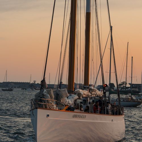 A sailboat yacht named &ldquo;ADIRONDACKY&rdquo; glides across the water at sunset, with sails furled and crew aboard.