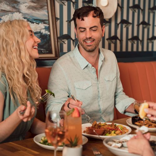 A couple enjoys dinner at a restaurant, smiling and eating with plates and drinks on the table in front of a fish-themed wall.